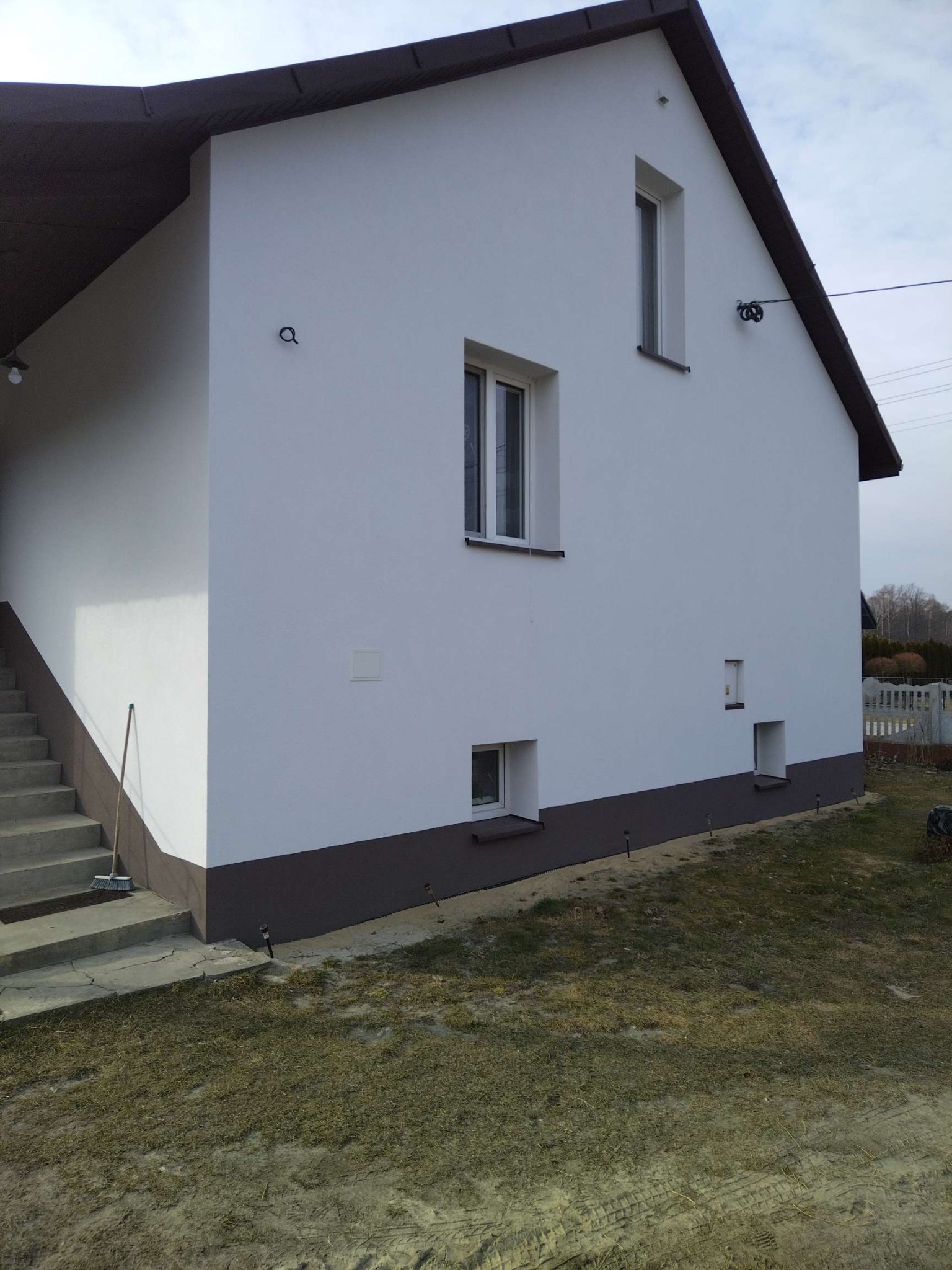 Boczny widok domu po renowacji elewacji. Biała ściana z brązowym cokołem, okna o różnych rozmiarach, schody wejściowe i fragment trawnika.