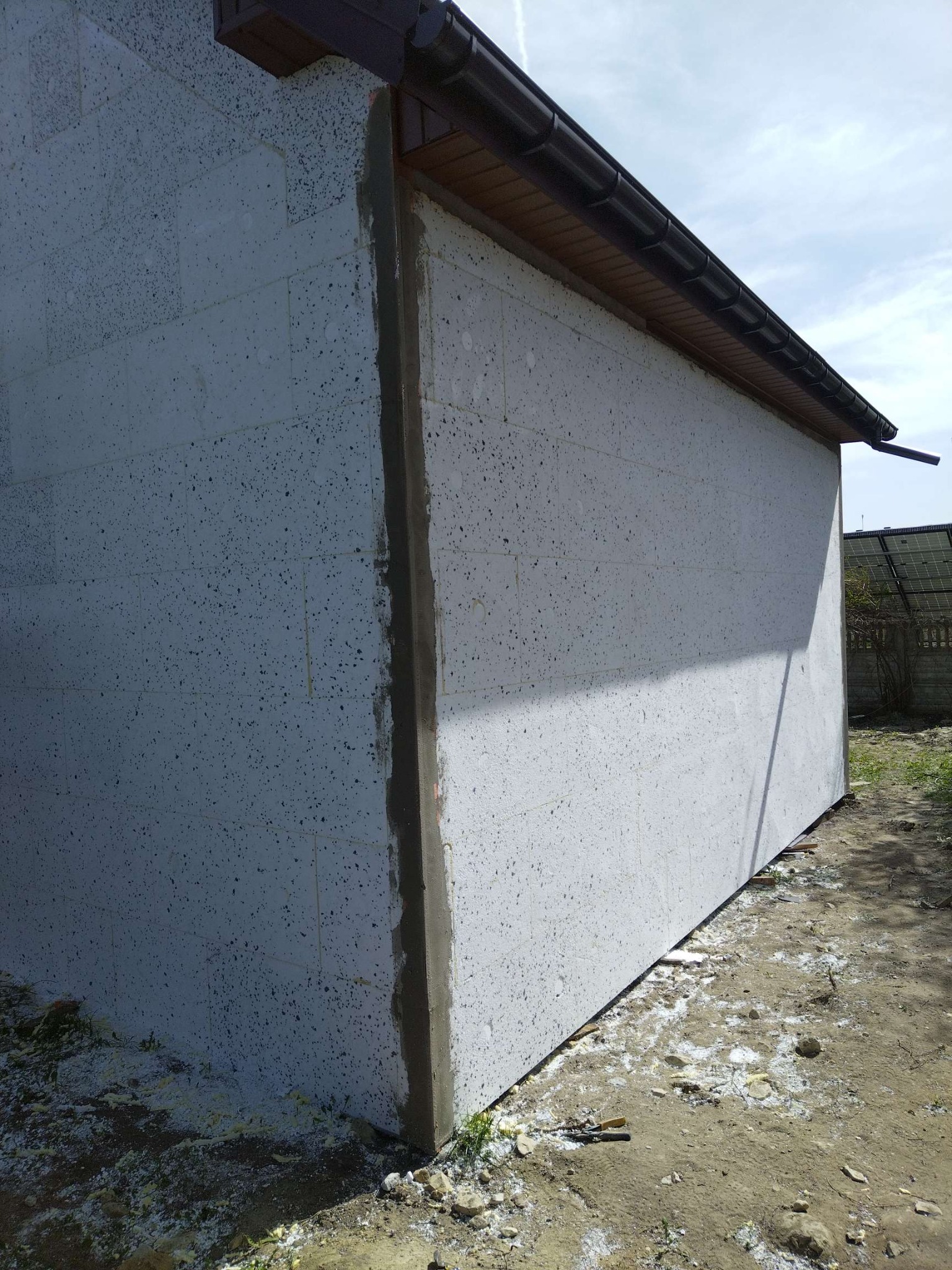 Ściana budynku w trakcie ocieplania styropianem, widoczne szpary wypełnione zaprawą. Na dole resztki materiału. Ujęcie z dołu, widoczny fragment dachu z rynną.