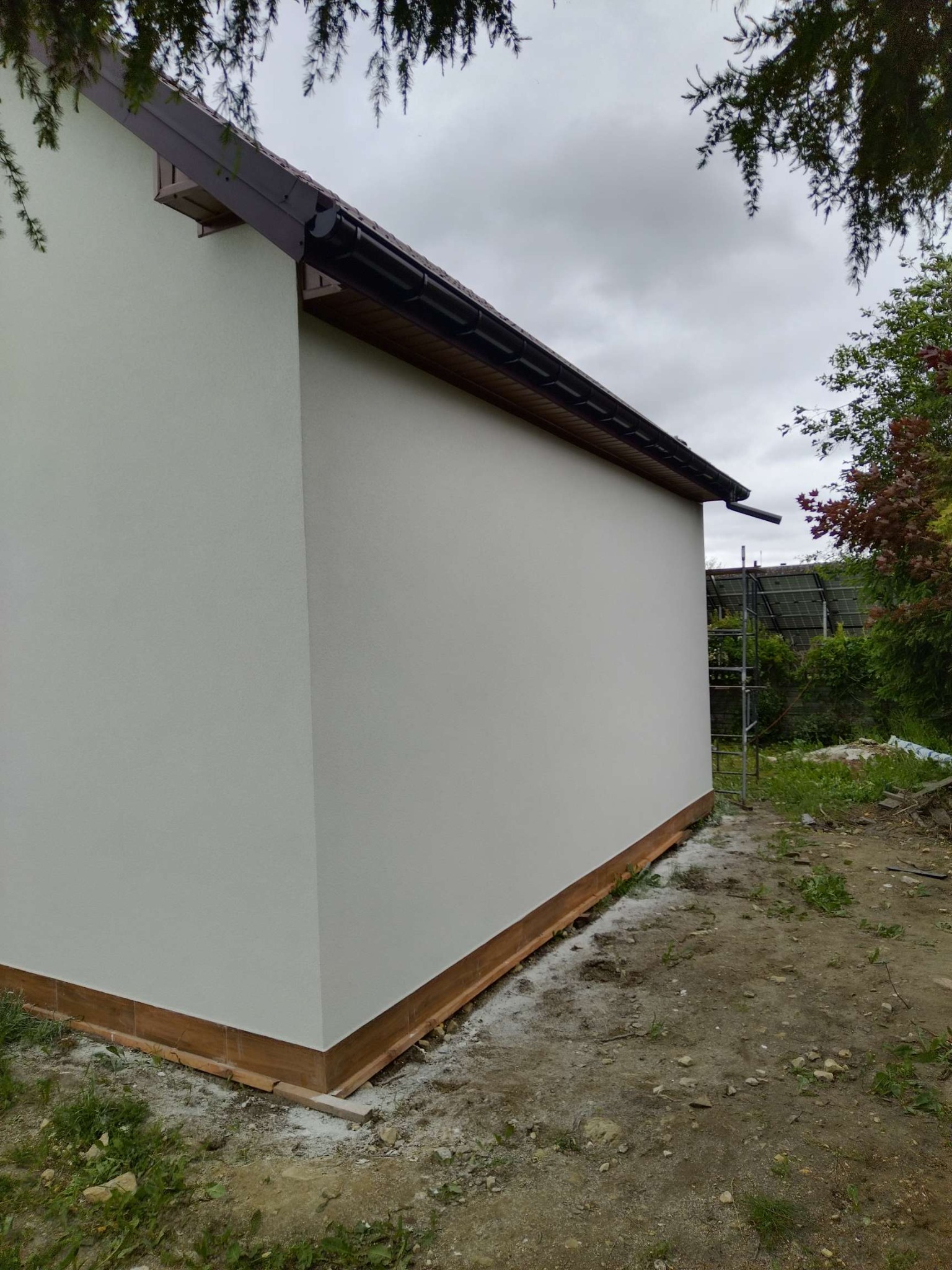 Świeżo otynkowana, biała ściana budynku z brązowym dachem i rynną. Widoczne rusztowanie z tyłu, zieleń i pochmurne niebo. Ukończony etap prac elewacyjnych.