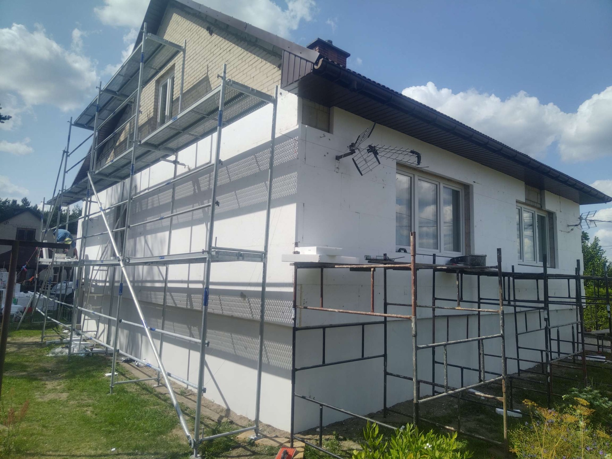 Dom w trakcie ocieplania, widoczne rusztowania i biały styropian na elewacji. Częściowo ocieplona ściana, z zamontowaną anteną. Prace w toku, słoneczny dzień.