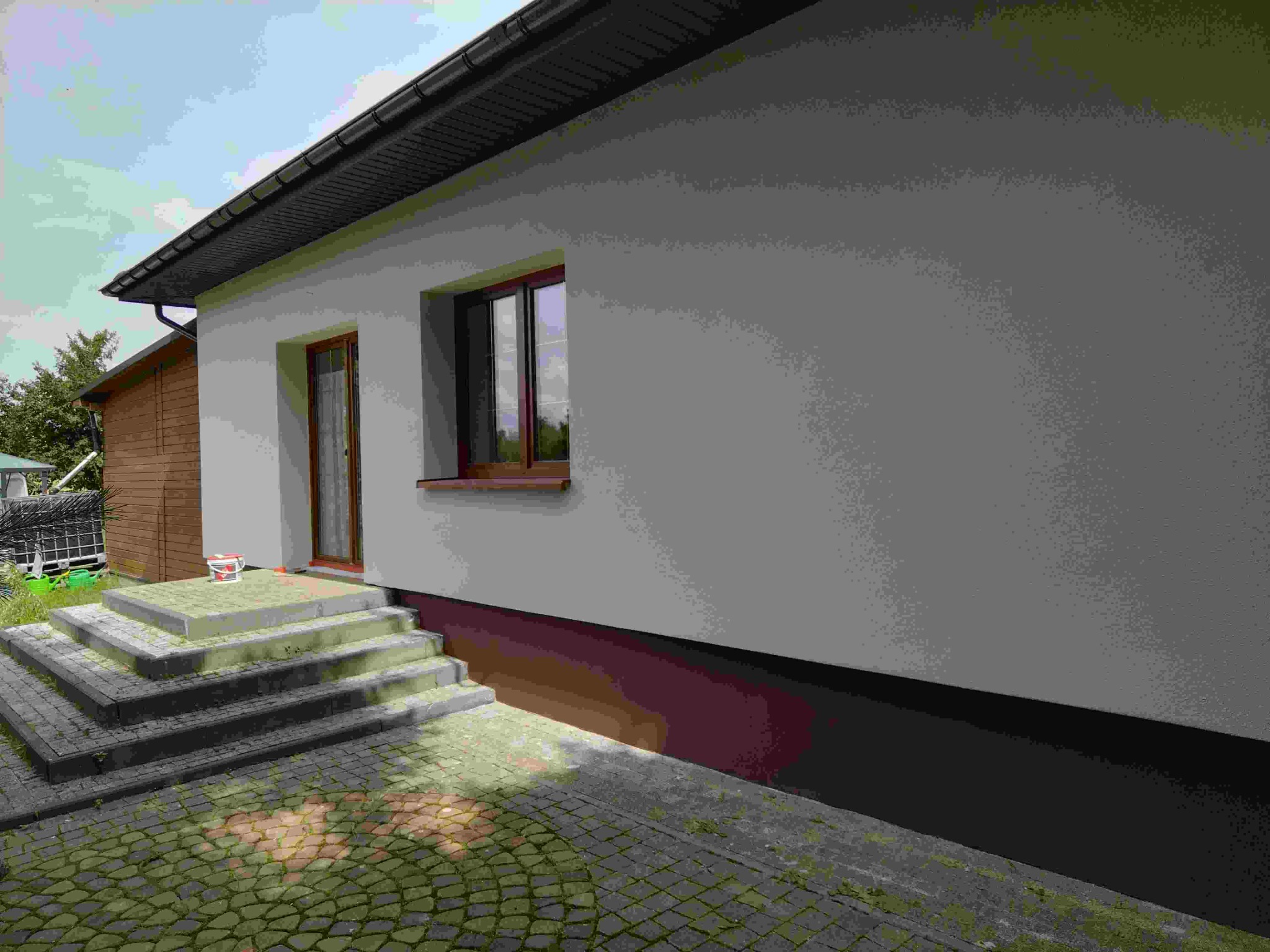 Świeżo otynkowana elewacja domu jednorodzinnego z brązowymi oknami i drzwiami. Widoczne schody wejściowe z kostki brukowej i wiadro z farbą.