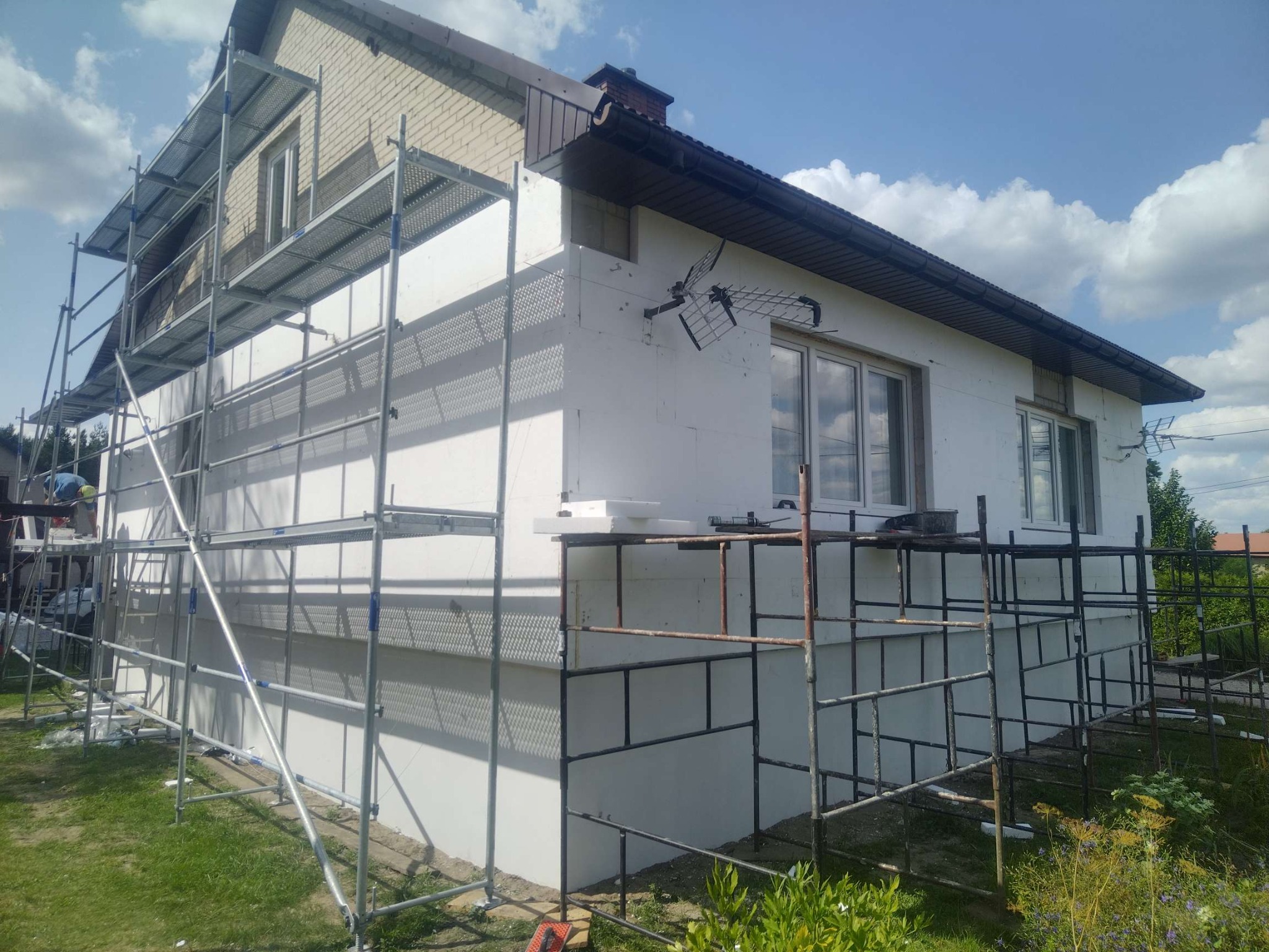 Dom jednorodzinny w trakcie ocieplania elewacji styropianem. Widoczne rusztowania, częściowo ocieplona ściana i antena telewizyjna na elewacji.