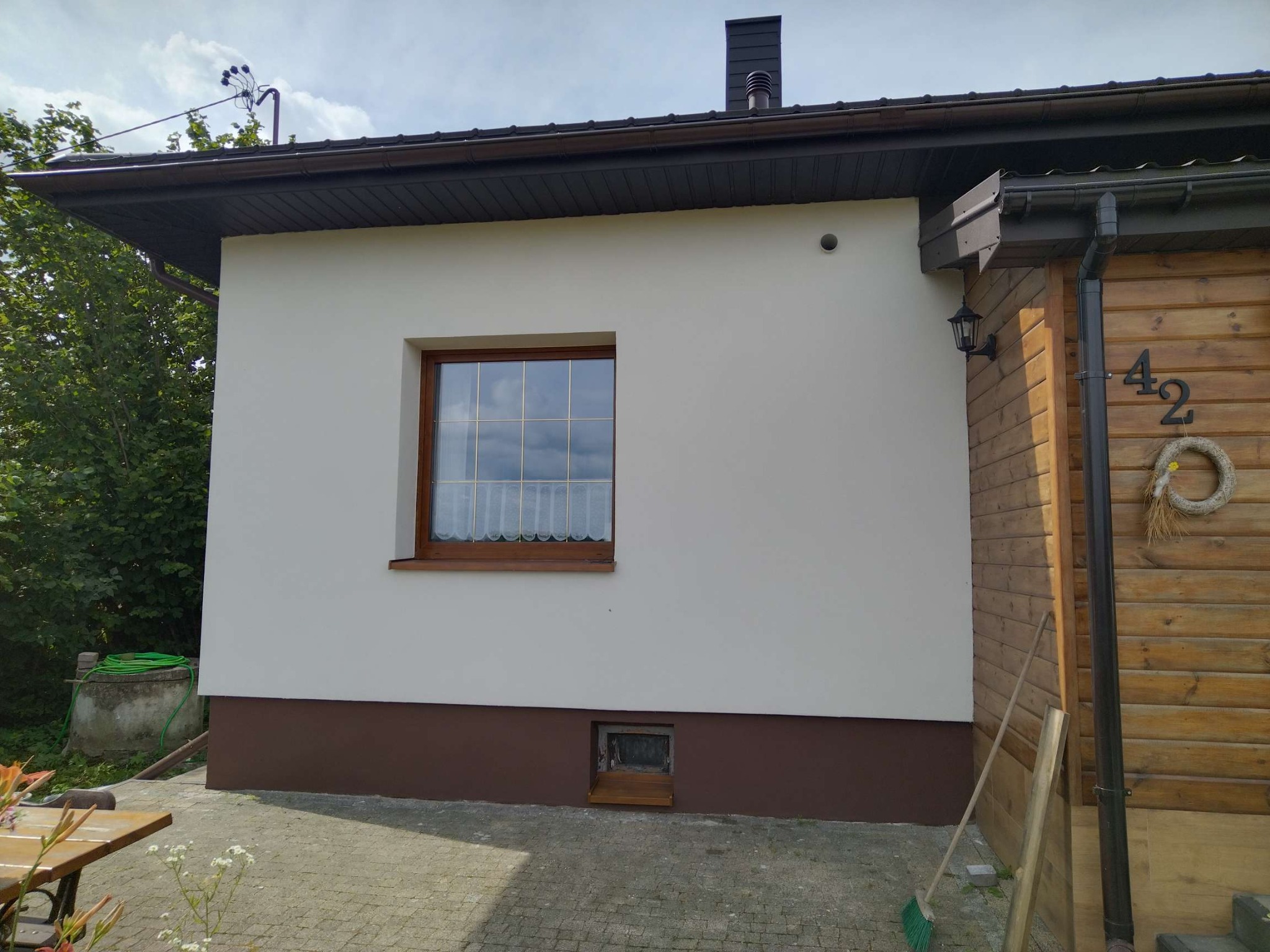 Ściana domu z oknem po renowacji elewacji. Jasny tynk, brązowe detale, drewniane wykończenia. Widoczny fragment numeru domu i narzędzia.
