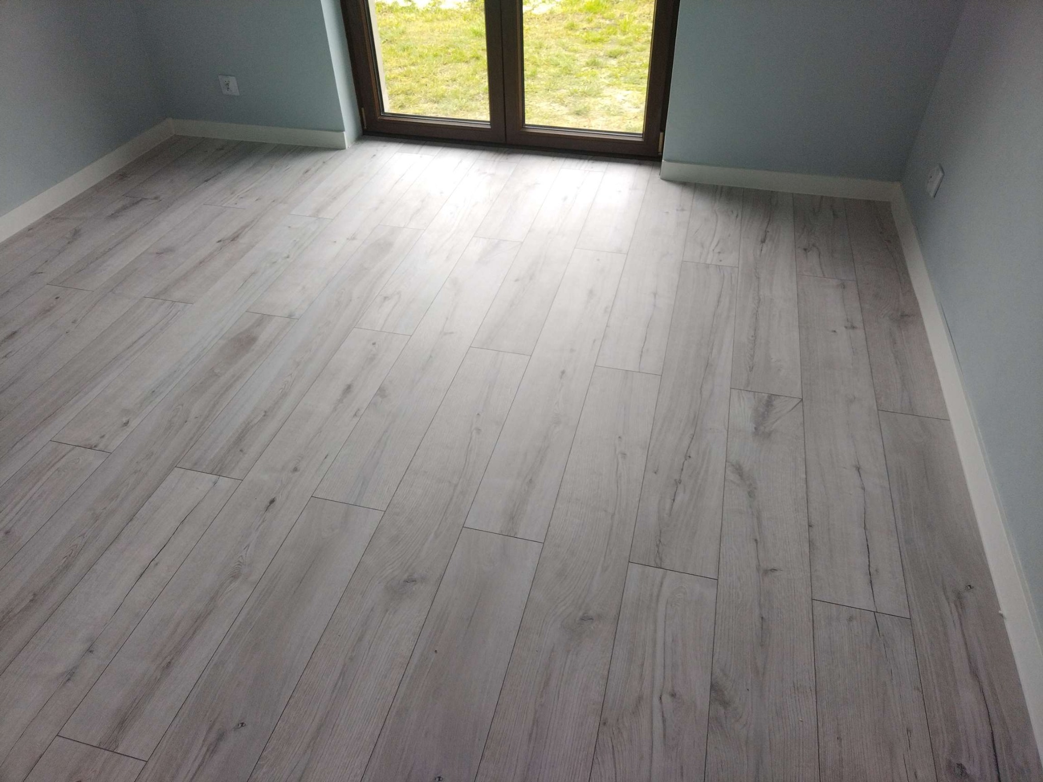 Jasnoszara podłoga z paneli ułożona w nowym pokoju z oknem balkonowym i niebieskimi ścianami. Widoczne listwy przypodłogowe i gniazdko.