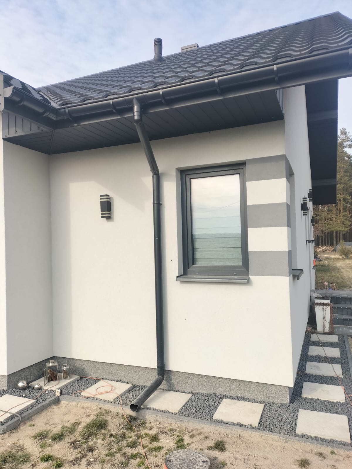 Boczny widok domu z białą elewacją, szarymi detalami okiennymi i pasami dekoracyjnymi. Widoczne rynny, oświetlenie zewnętrzne i chodnik z płyt.