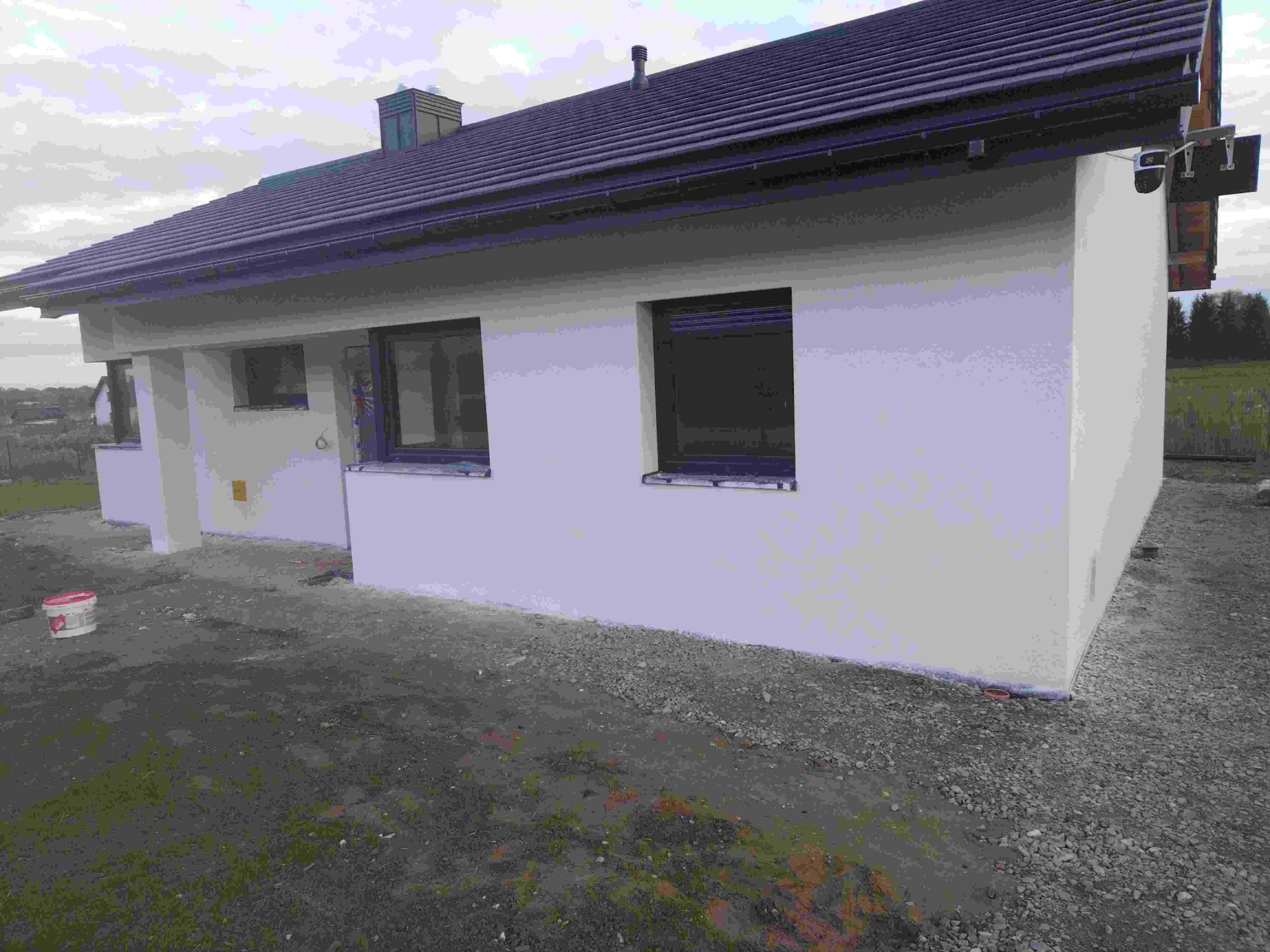 Otynkowany dom jednorodzinny z ciemnym dachem i oknami. Widoczny fragment elewacji bocznej i otoczenie domu. Na ziemi stoi wiadro z farbą.