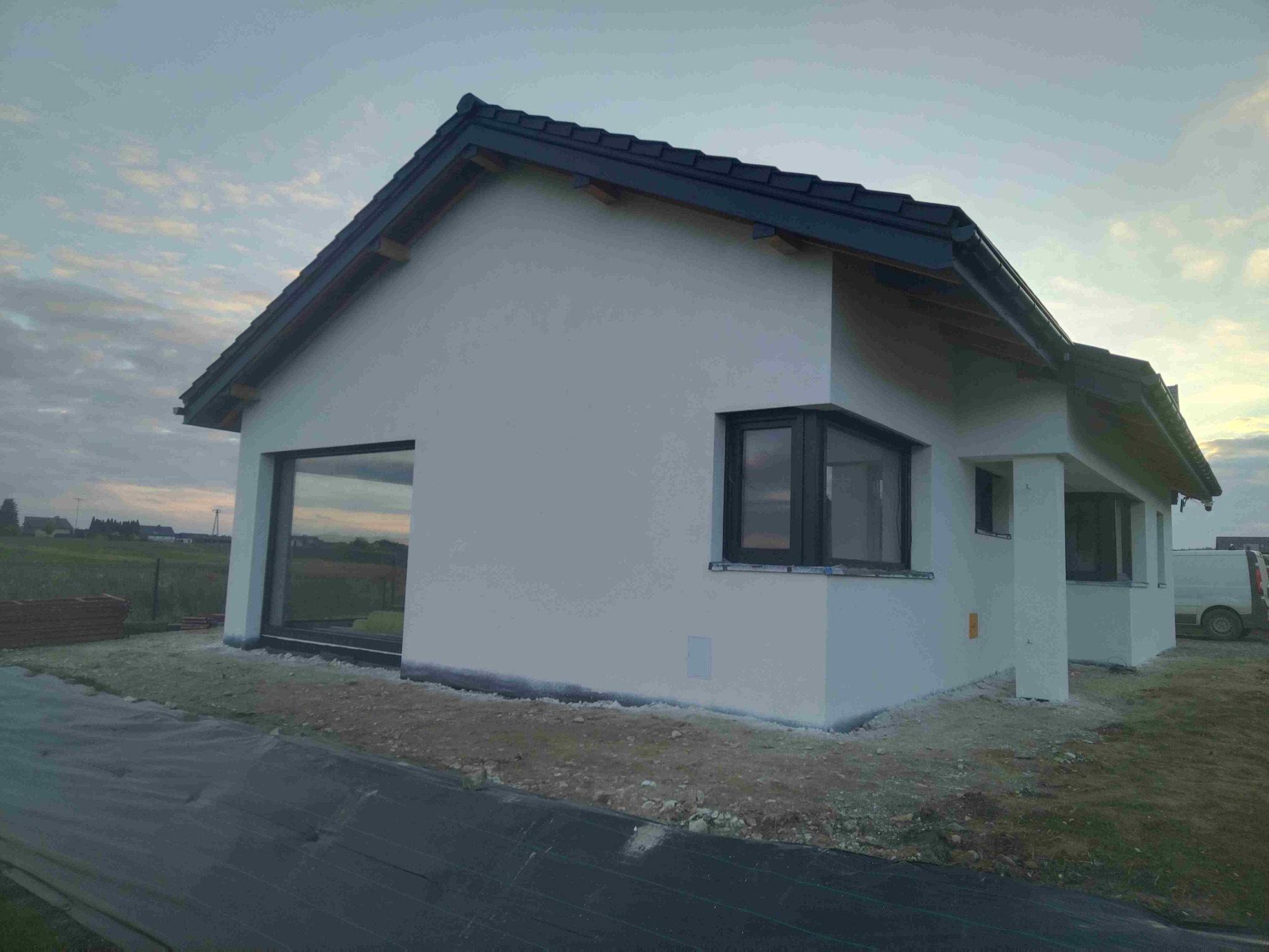 Nowoczesny dom jednorodzinny z białymi ścianami i ciemną dachówką, widoczny z zewnątrz, z dużym oknem i elementami wykończeniowymi w kolorze antracytowym.