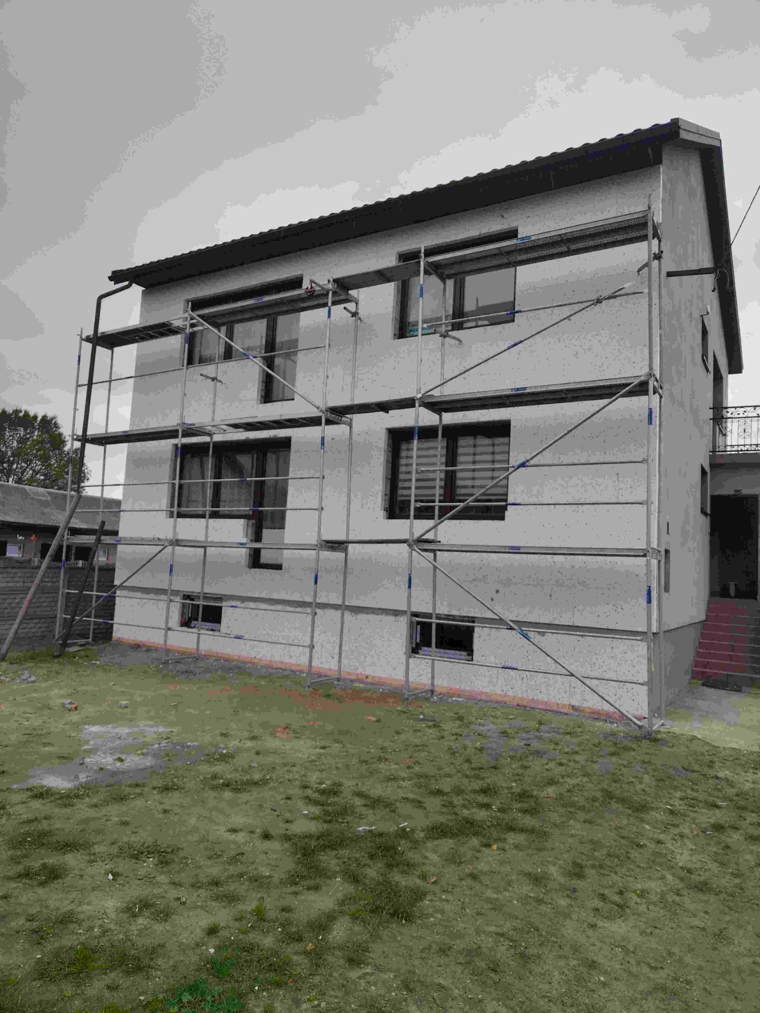 Dom jednorodzinny w trakcie ocieplania, z widocznymi rusztowaniami i szarym tynkiem. Okna z roletami. Widok z poziomu gruntu na elewację.