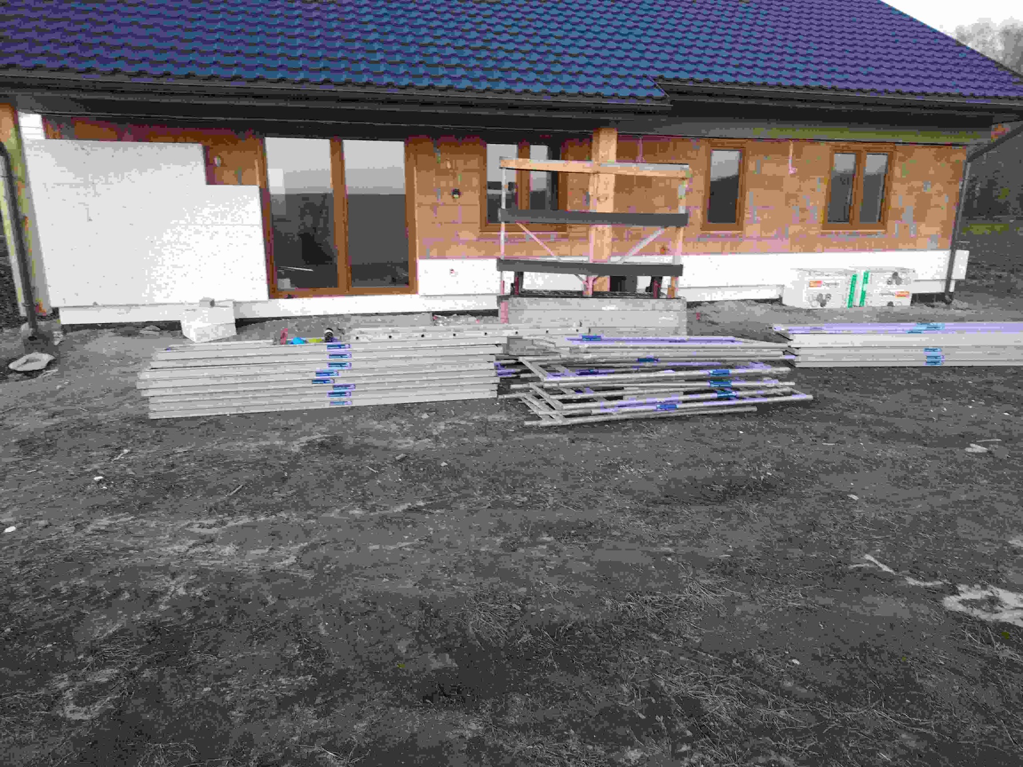 Dom w trakcie ocieplania: fragment elewacji z oknami, częściowo pokryty styropianem, rusztowania i materiały budowlane na ziemi. Widoczny niebieski dach z dachówki.