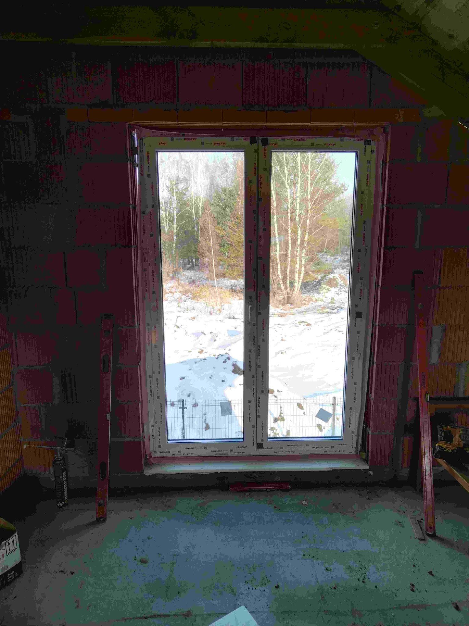 Montaż okna w surowym stanie: białe okno PCV w otworze z czerwonej cegły, poziomice po bokach, widok na zimowy krajobraz z brzozami. Etap prac wykończeniowych.