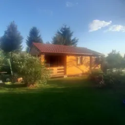 Drewniany domek letniskowy z czerwonym dachem z dachówki bitumicznej, otoczony zielenią drzew i trawnikiem w słoneczny dzień.