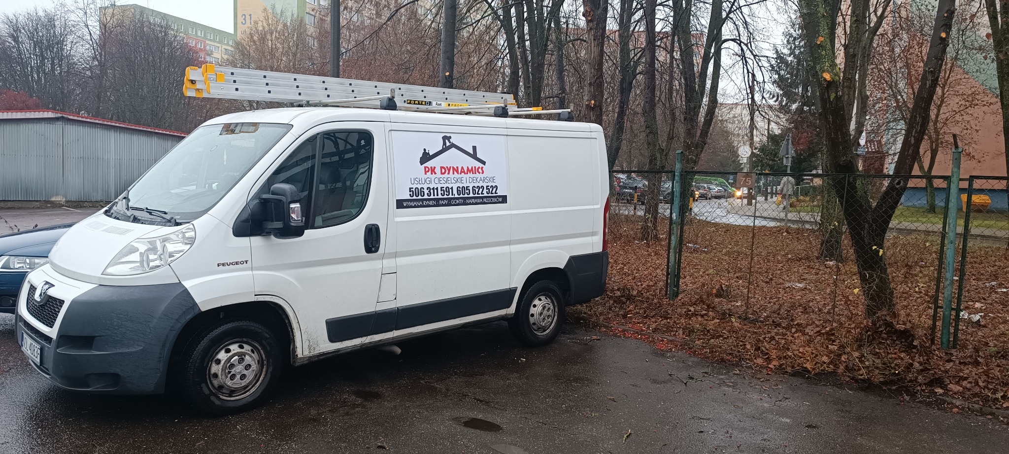 Biały samochód dostawczy firmy dekarskiej PK Dynamics z drabiną na dachu, zaparkowany na parkingu z jesiennymi liśćmi, widoczne logo firmy z sylwetką dekarza i numerami telefonów.