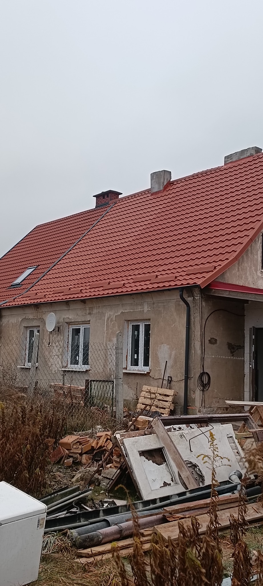 Dom z nowym, czerwonym dachem z dachówki ceramicznej, widoczne nowe okna, nieotynkowana elewacja i gruz budowlany na posesji.