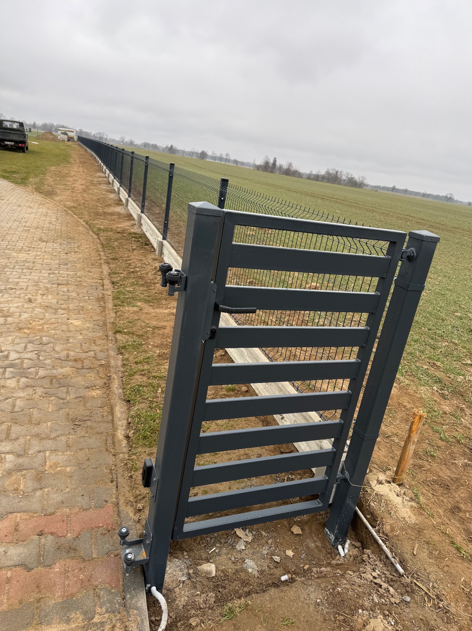 Nowoczesna, grafitowa brama wjazdowa z poziomymi szczeblami, stanowiąca część ogrodzenia panelowego na betonowej podmurówce. Widok na pole i pochmurne niebo.