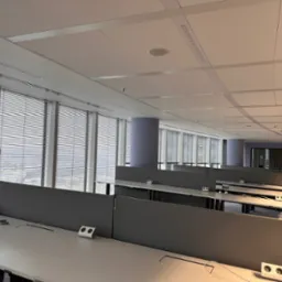 Nowoczesne biuro w wysokim budynku z rzędami białych biurek z szarymi przegrodami i wbudowanymi gniazdkami, widok na miasto przez żaluzje okienne.