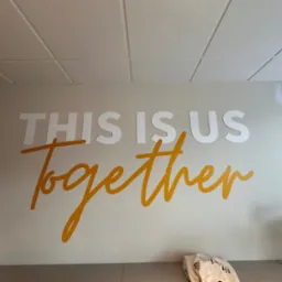 Biała ściana biura z napisem 'THIS IS US Together' w kolorze białym i złotym, podświetlonym przez światło padające z sufitu kasetonowego.