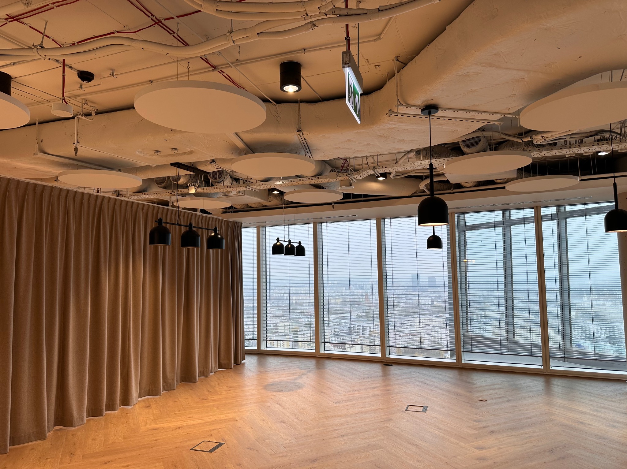 Wnętrze biura z dużymi oknami z widokiem na miasto, drewnianą podłogą w jodełkę, beżowymi zasłonami i surowym sufitem z widocznymi instalacjami.