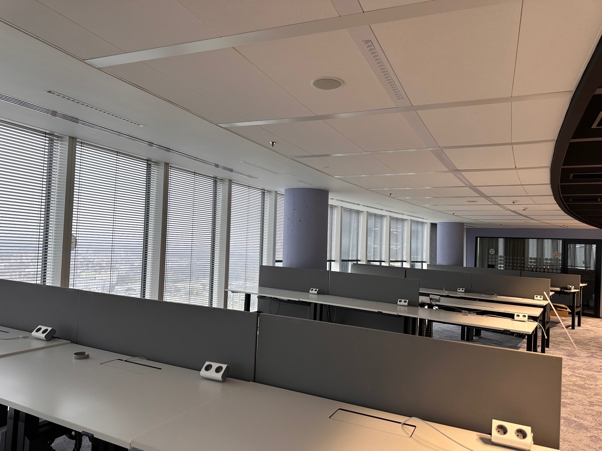 Nowoczesne biuro w wysokim budynku z rzędami białych biurek z szarymi przegrodami i wbudowanymi gniazdkami, widok na miasto przez żaluzje okienne.