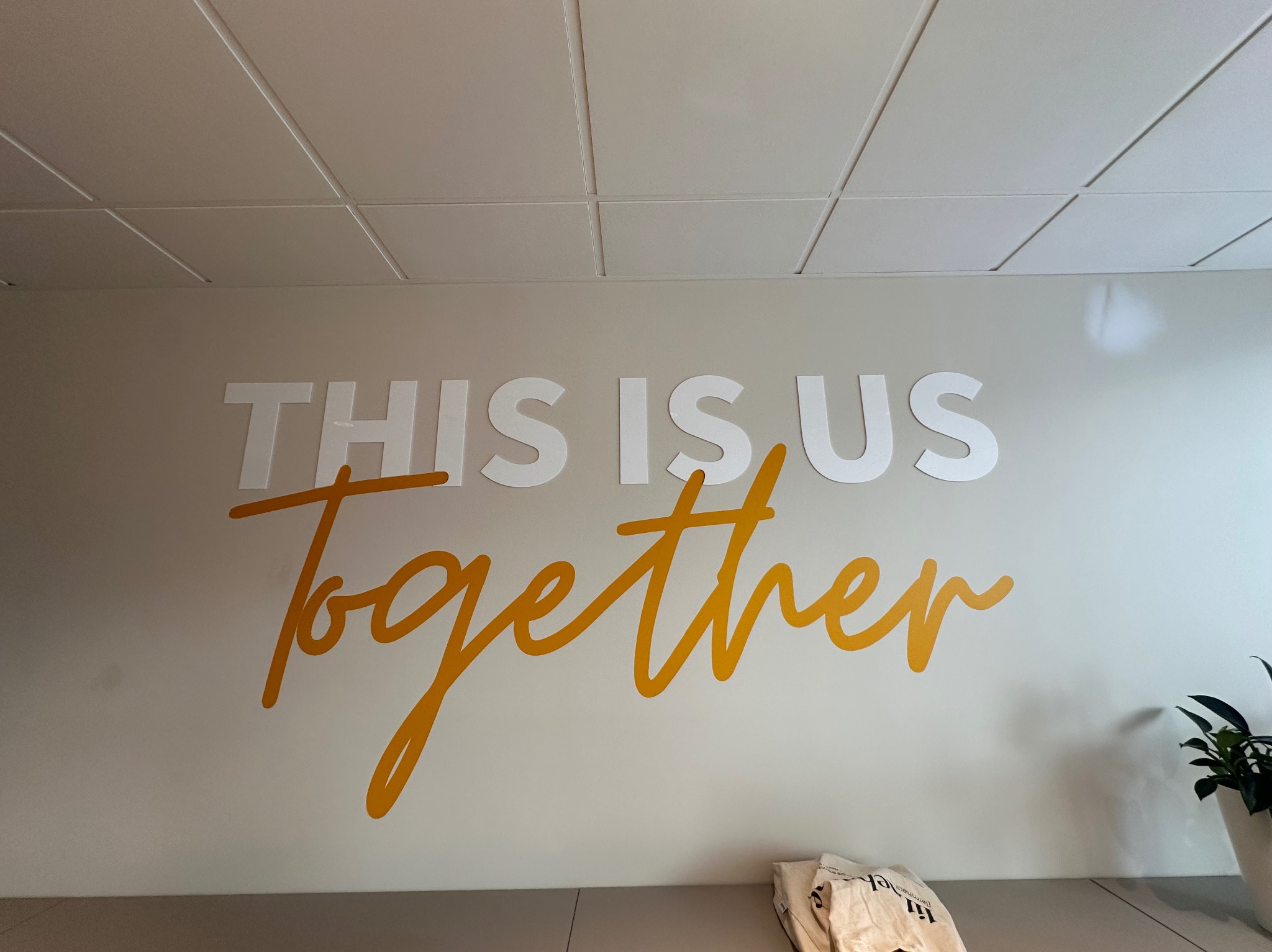 Biała ściana biura z napisem 'THIS IS US Together' w kolorze białym i złotym, podświetlonym przez światło padające z sufitu kasetonowego.