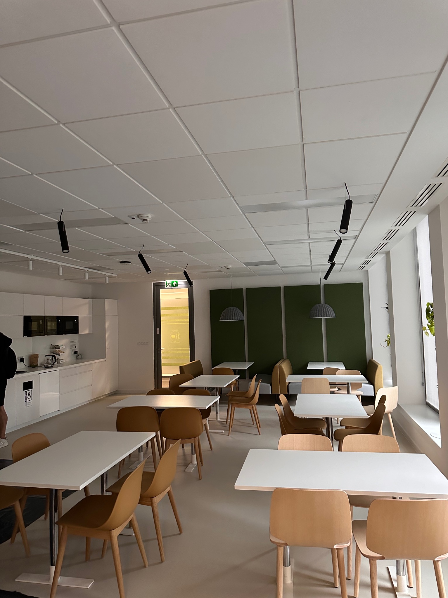 Jasne biuro z białymi stołami, drewnianymi krzesłami, aneksem kuchennym i panelami akustycznymi w odcieniach zieleni.
