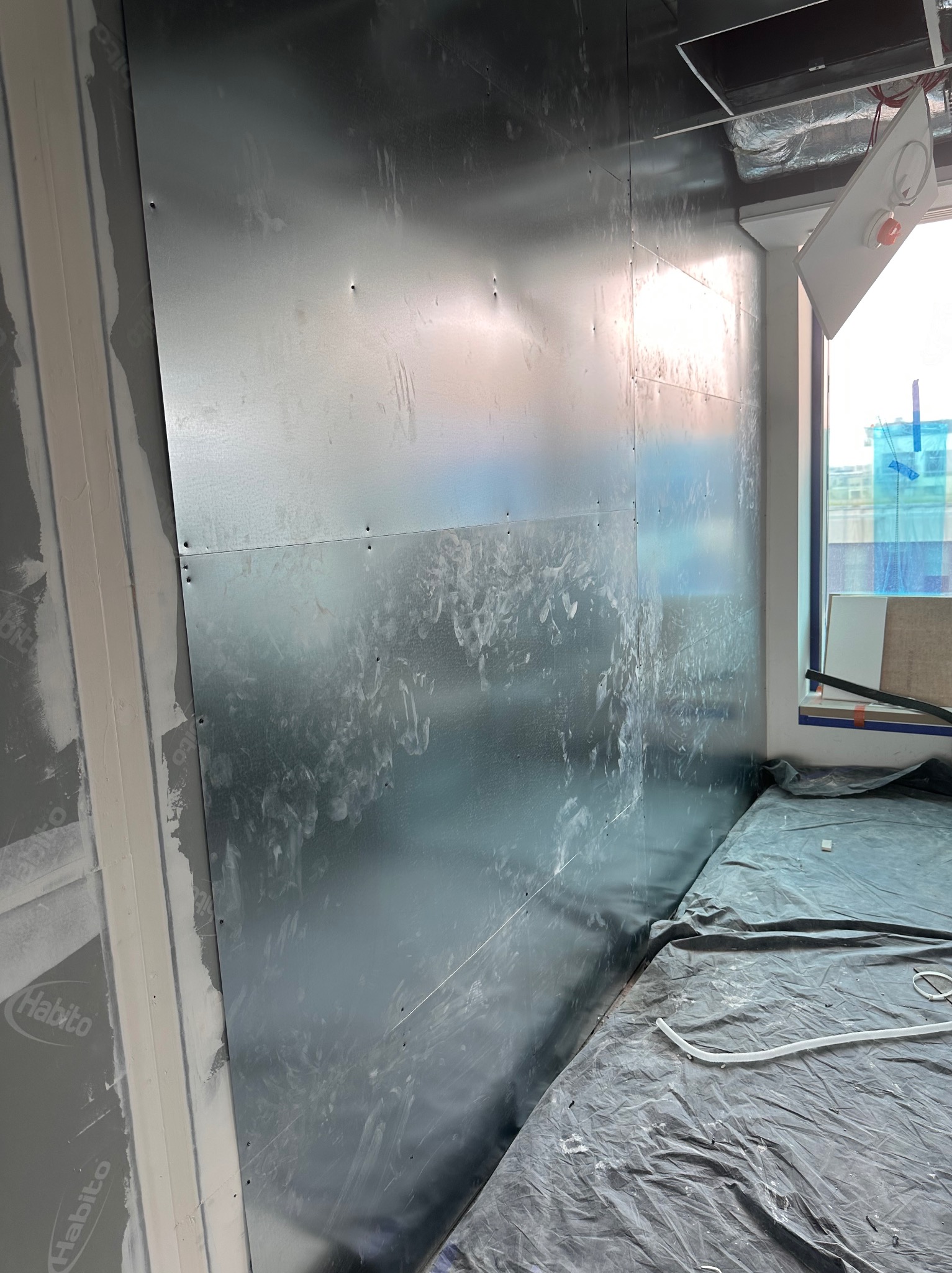 Montaż płyt kartonowo-gipsowych na ścianie, widoczne ślady po szpachlowaniu, zabezpieczona folią podłoga, surowy stan wykończenia wnętrza.