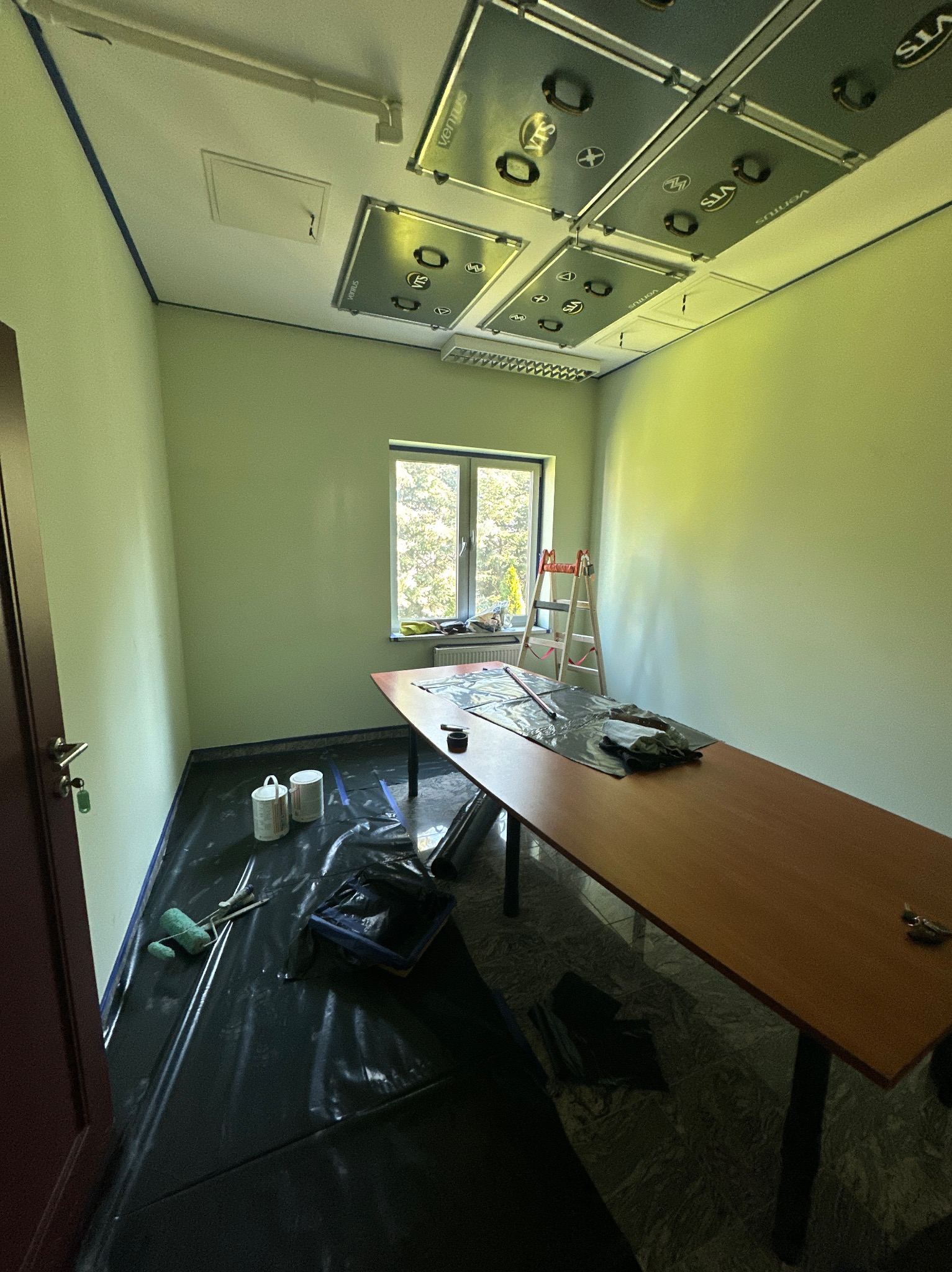 Biuro w trakcie odświeżania: świeżo pomalowane ściany w odcieniach zieleni, zabezpieczona folią podłoga, drabina oparta o ścianę przy oknie, widoczne puszki farby i narzędzia malarskie.