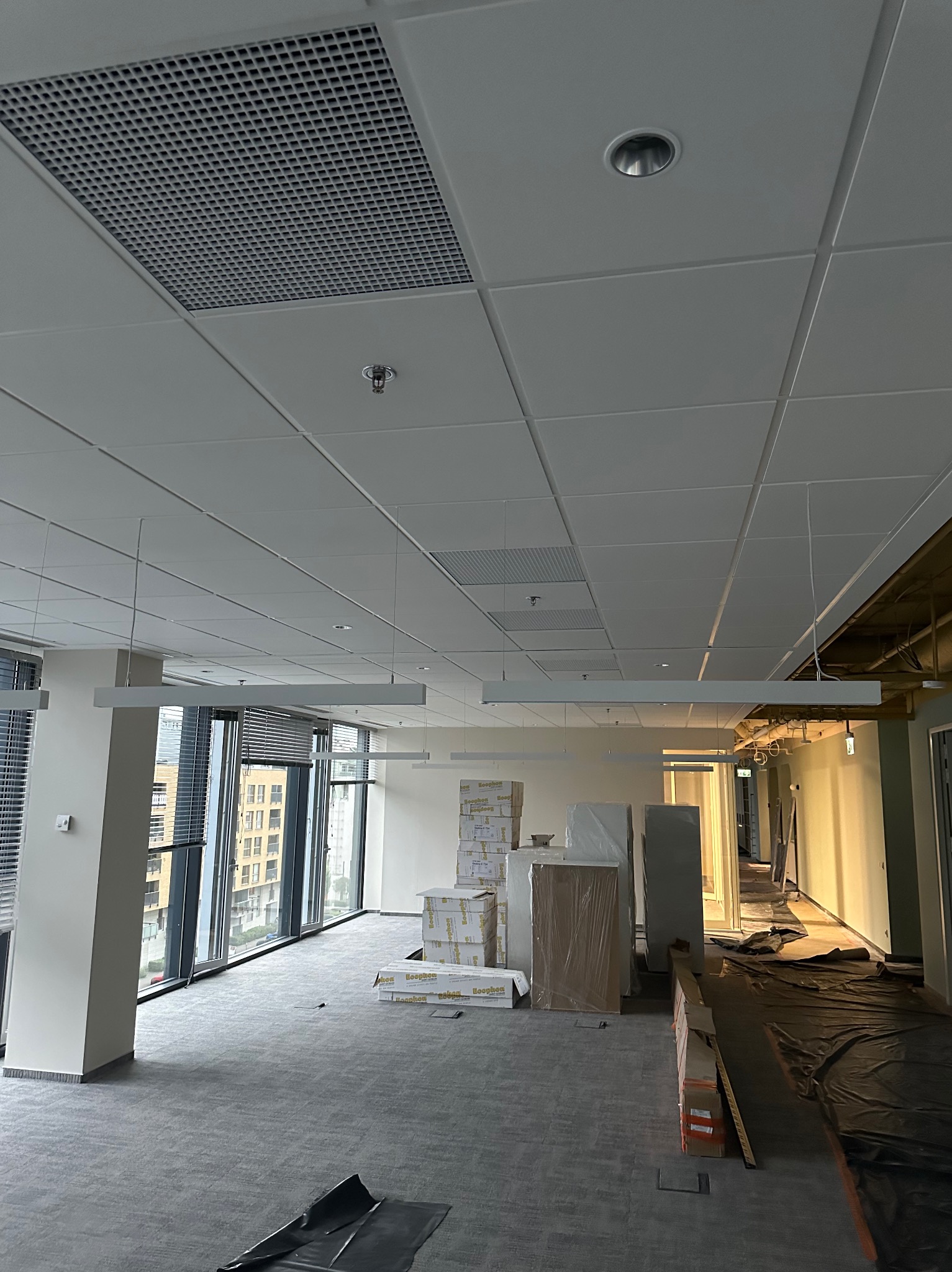 Przestronne biuro w trakcie prac wykończeniowych: widok na sufit podwieszany z kratką wentylacyjną i lampami, podłoga wyłożona wykładziną dywanową, okna z żaluzjami, materiały budowlane...