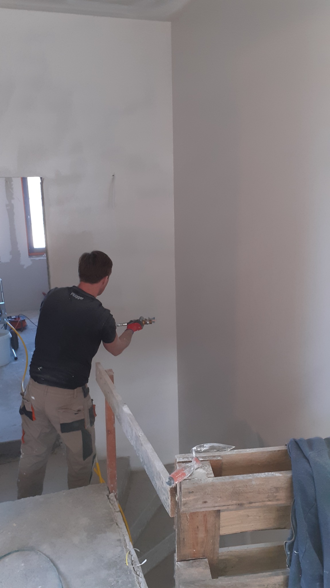 Pracownik w okularach ochronnych aplikuje białą masę na ścianę za pomocą pistoletu natryskowego w pomieszczeniu w trakcie wykańczania.