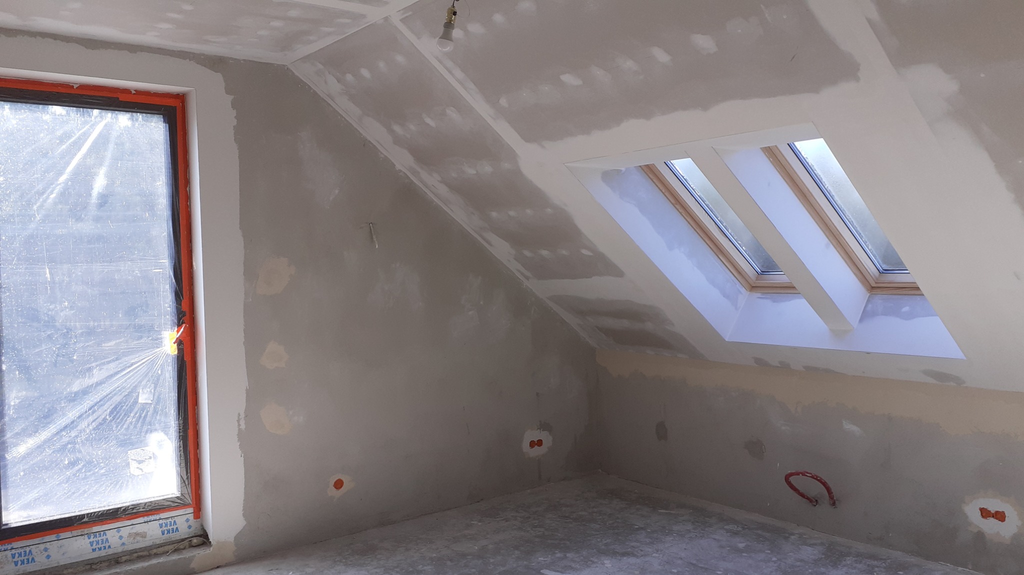 Pokój na poddaszu w trakcie szpachlowania ścian i sufitu, widoczne okno dachowe, okno pionowe zabezpieczone folią, niedokończone gładzie i instalacja elektryczna.