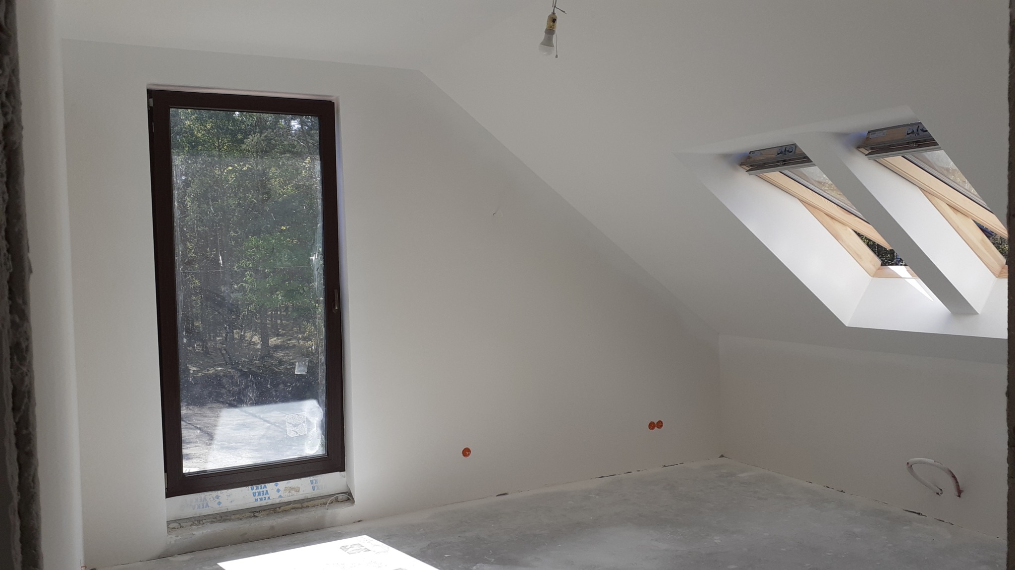 Poddasze po pracach przygotowawczych: białe ściany, dwa okna dachowe z drewnianą ramą, brązowe okno pionowe, widoczne gniazdka elektryczne i wystający kabel.