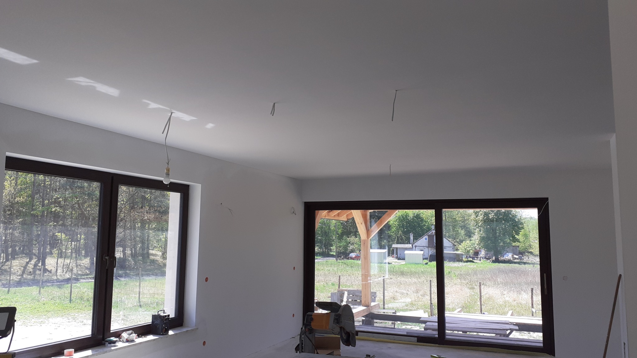Wnętrze w trakcie prac wykończeniowych: gładkie, białe ściany i sufit, duże okna z widokiem na zewnątrz, widoczne przewody elektryczne i narzędzia na podłodze.