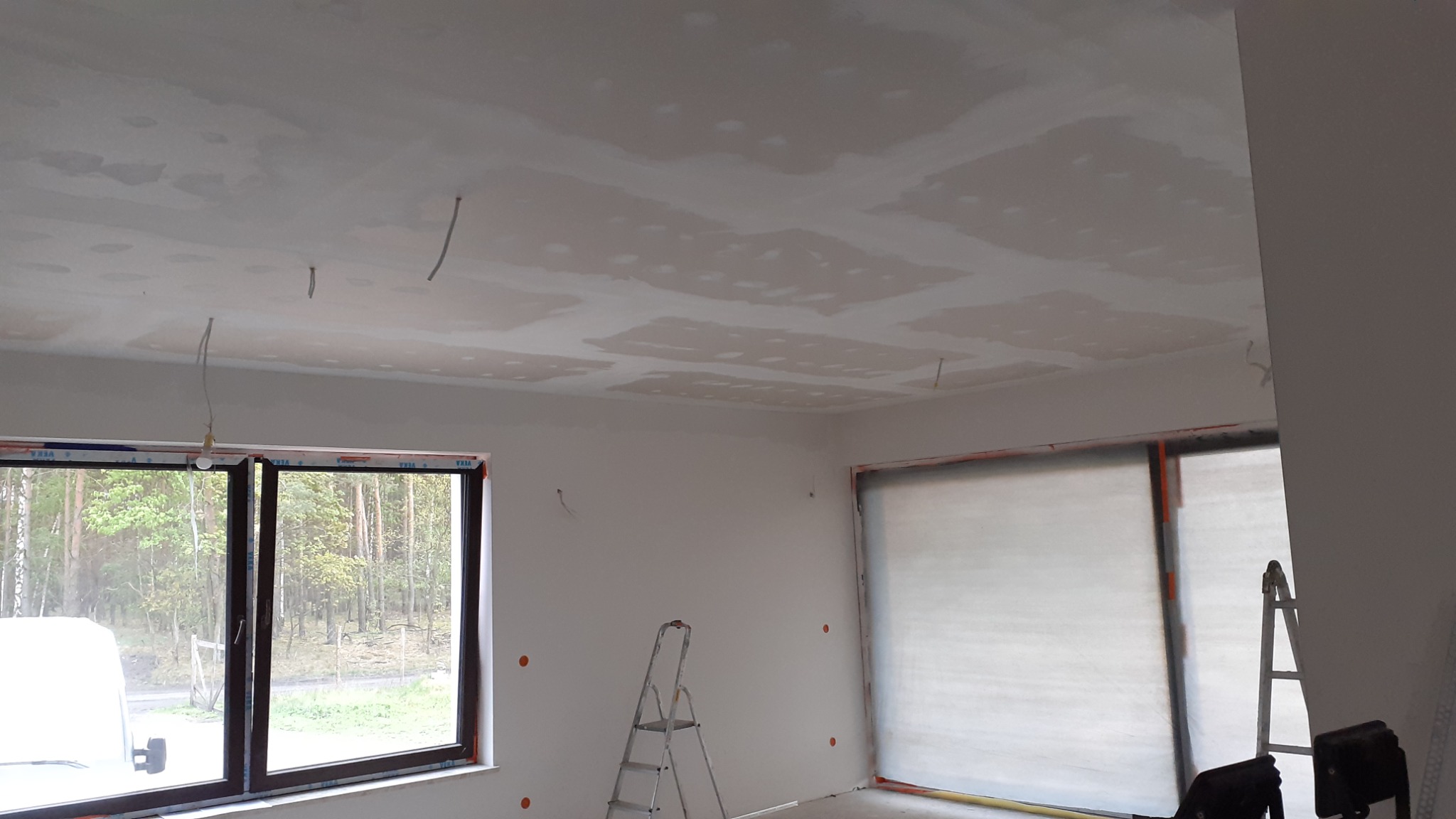 Pokój w trakcie prac: sufit z widocznymi łączeniami płyt gipsowo-kartonowych przed szlifowaniem, widoczne wystające przewody elektryczne, okna z widokiem na drzewa, drabina w rogu pomieszczenia.
