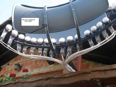 Duża antena satelitarna typu offset z wieloma konwerterami LNB zamontowana na dachu budynku z czerwonej cegły, widoczne kable i czerwone kwiaty w doniczce na pierwszym planie.