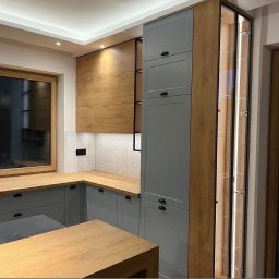 Harmony Kitchens - Szara zabudowa kuchenna z drewnianymi akcentami, podświetlana witryna narożna i płytki ułożone w jodełkę na ścianie nad blatem.