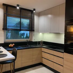 Nowoczesna kuchnia w kształcie litery L z drewnianymi frontami szafek, czarnym blatem i roletami w oknie, oświetlona punktowymi lampami sufitowymi i listwą LED pod szafkami wiszącymi.