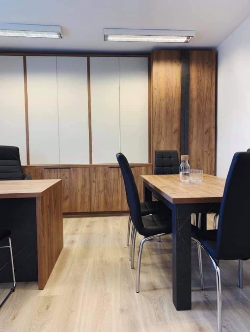 Biuro z szafami na wymiar z białymi frontami i drewnianymi elementami, biurko z blatem drewnianym i czarną podstawą, stół z czarnymi nogami i drewnianym blatem, czarne krzesła na srebrnych nogach...