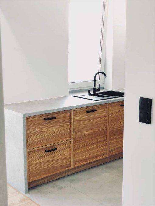 Nowoczesna zabudowa kuchenna z drewnianymi frontami, szarym blatem i czarną armaturą, minimalistyczny design, widok z perspektywy wejścia.