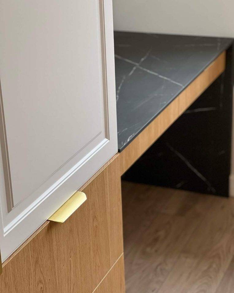 Fragment zabudowy kuchennej: białe fronty szafek z frezowanymi ramkami, złote uchwyty, blat z czarnego marmuru z białym żyłowaniem, drewnopodobne wykończenie korpusu szafek.