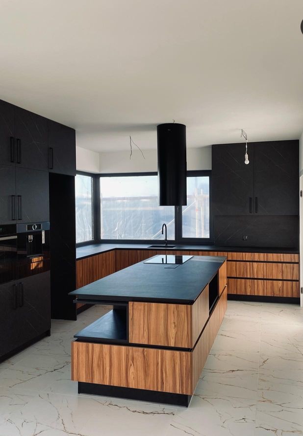 Nowoczesna kuchnia z wyspą, fronty szafek z ciemnego drewna z czarnymi akcentami, czarny okap cylindryczny, marmurowe płytki podłogowe, widoczne niedokończone prace wykończeniowe.