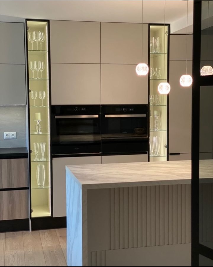 Nowoczesna kuchnia z wyspą, fronty w kolorze beżowym, zabudowane piekarniki, podświetlane przeszklone witryny z kieliszkami, marmurowy blat wyspy.