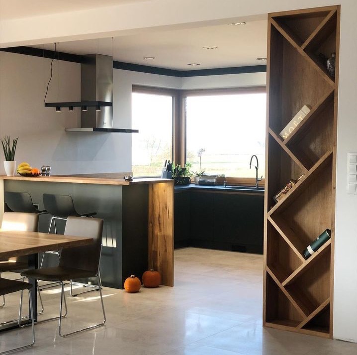 Nowoczesna kuchnia z wyspą, ciemnymi szafkami, drewnianym blatem i dekoracyjnym regałem na wino z butelkami Moet, oświetlona naturalnym światłem z dużego okna, z dyniami jako elementem dekoracyjnym...