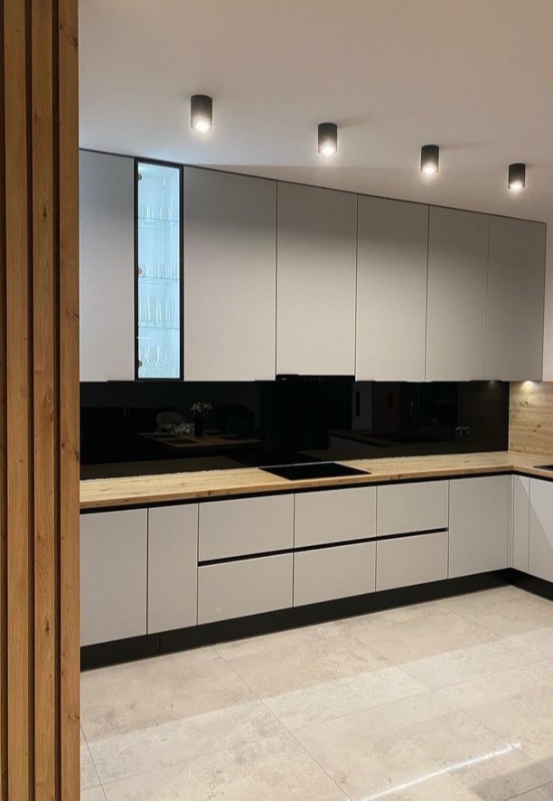 Nowoczesna kuchnia w odcieniach szarości z drewnianym blatem i czarnym panelem nad blatem, oświetlona punktowymi lampami sufitowymi i z przeszkloną szafką eksponującą szkło.