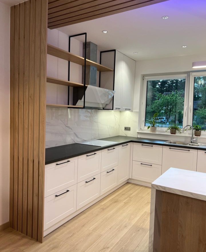Nowoczesna kuchnia w bieli z czarnymi akcentami, drewniane półki i panele dekoracyjne, widok na zieleń za oknem.