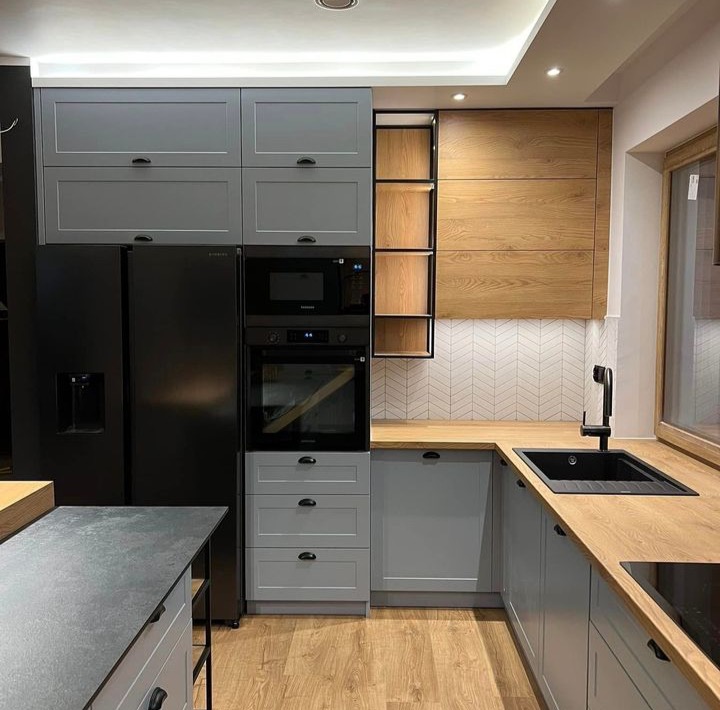 Nowoczesna kuchnia w odcieniach szarości i drewna z czarnymi akcentami, widok na zabudowę kuchenną z szafkami górnymi, dolnymi, lodówką, piekarnikiem i mikrofalówką, drewniany blat i białe płytki...