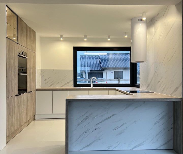 Nowoczesna kuchnia z wyspą, fronty szafek w odcieniach drewna i bieli, marmurowe wykończenie ściany, okno z widokiem na dom.