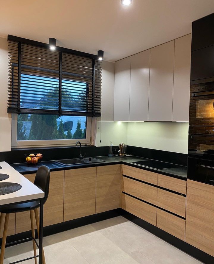 Nowoczesna kuchnia w kształcie litery L z drewnianymi frontami szafek, czarnym blatem i roletami w oknie, oświetlona punktowymi lampami sufitowymi i listwą LED pod szafkami wiszącymi.