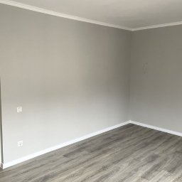 Andrzej Samerdak - Pomalowane na szaro wnętrze pokoju z szarymi panelami podłogowymi i białymi listwami przypodłogowymi oraz sufitowymi, widoczne gniazdko elektryczne i włącznik światła.