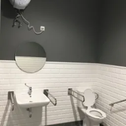 Wykończona łazienka z szarymi płytkami podłogowymi, białymi płytkami ściennymi typu metro, umywalką, toaletą z uchwytami dla niepełnosprawnych i okrągłym lustrem na szarej ścianie, nad umywalką...