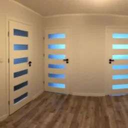 Wnętrze po remoncie: trzy białe drzwi z poziomymi, niebieskimi przeszkleniami, brązowe panele podłogowe, białe ściany i grzejnik.