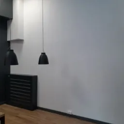 Wnętrze po remoncie: świeżo pomalowana biała ściana, drewnopodobne panele na podłodze, czarny grzejnik i dwie czarne lampy wiszące.